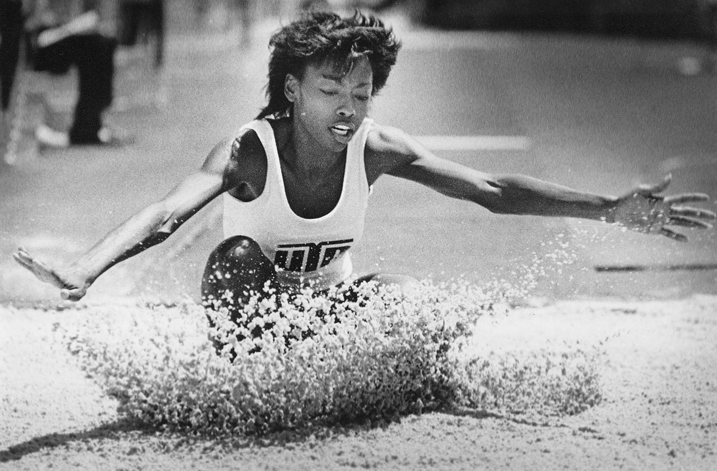 Black and white photo of a track meet with a track member jumping and landing in a pit of sand.