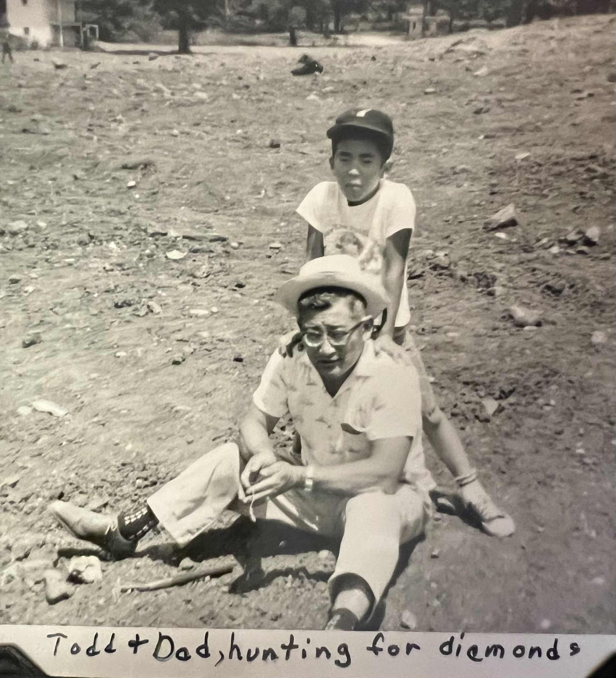 Hiro Hayataka sitting on the dirt ground, his son, Todd, leaning on his shoulders. Text reads ''Todd and Dad, hunting for diamonds'.