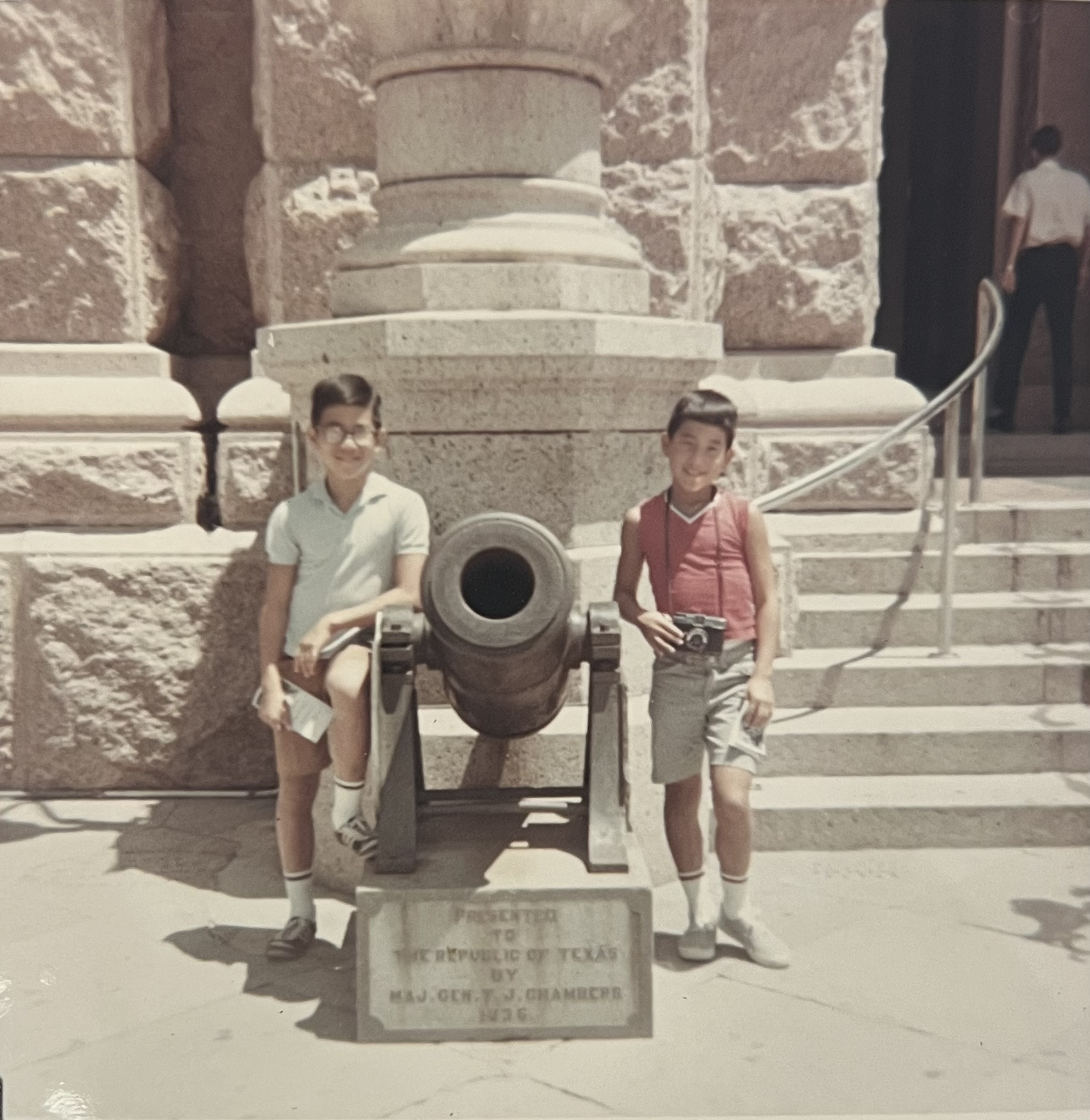 Todd and Glenn Hayataka posing in front of the Texas State Capitol, between them is a cannon with the text 'Presented to the Republic of Texas by Maj. Gen. T. J. Chambers 1836'. 