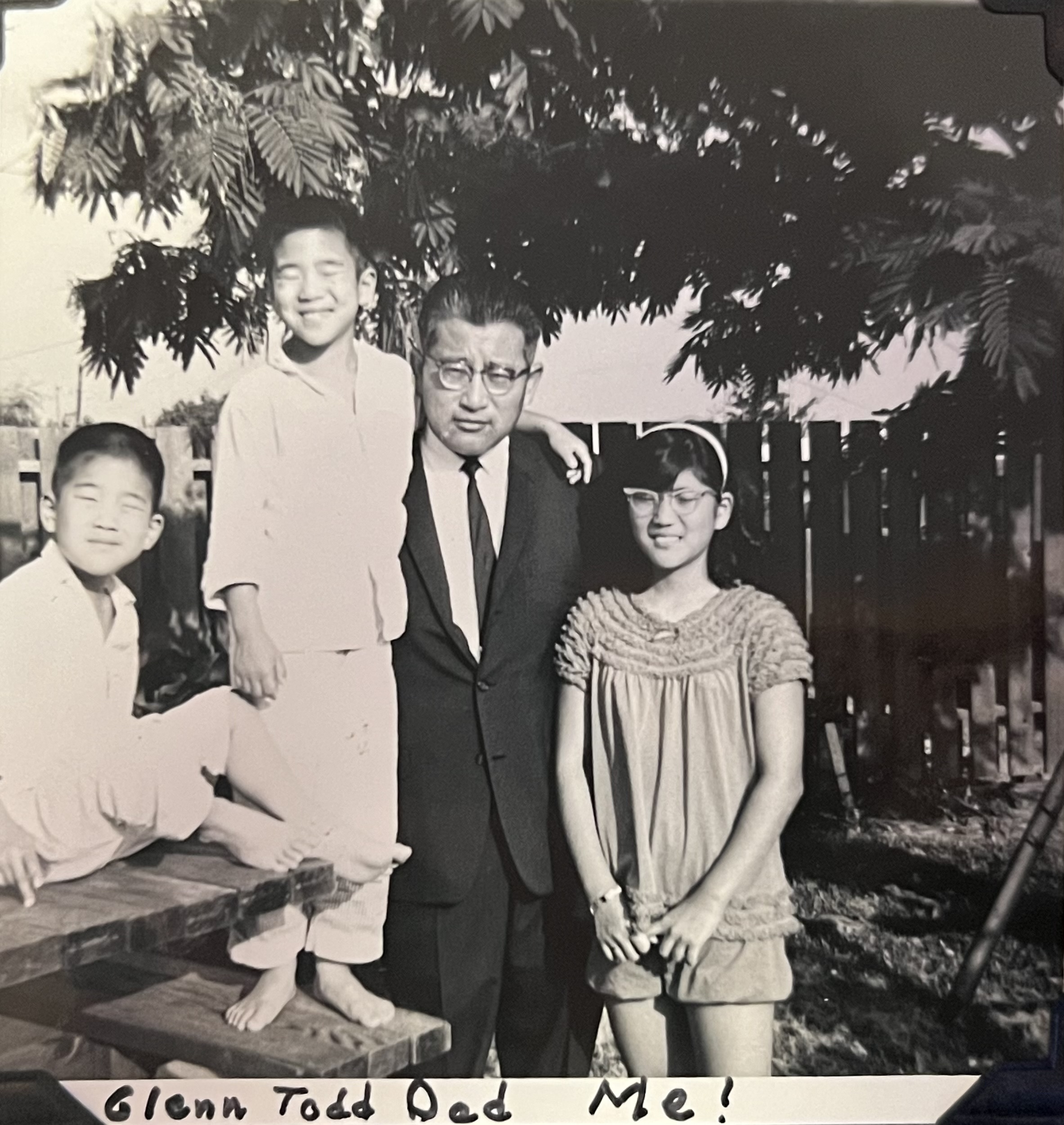 Three Japanese American children wearing 1960s pajamas, two boys and one girl, and a Japanese American man in a suit and glasses. They are in a yard in front of a fence. Text on the photo reads 'Glenn Todd Dad Me!'