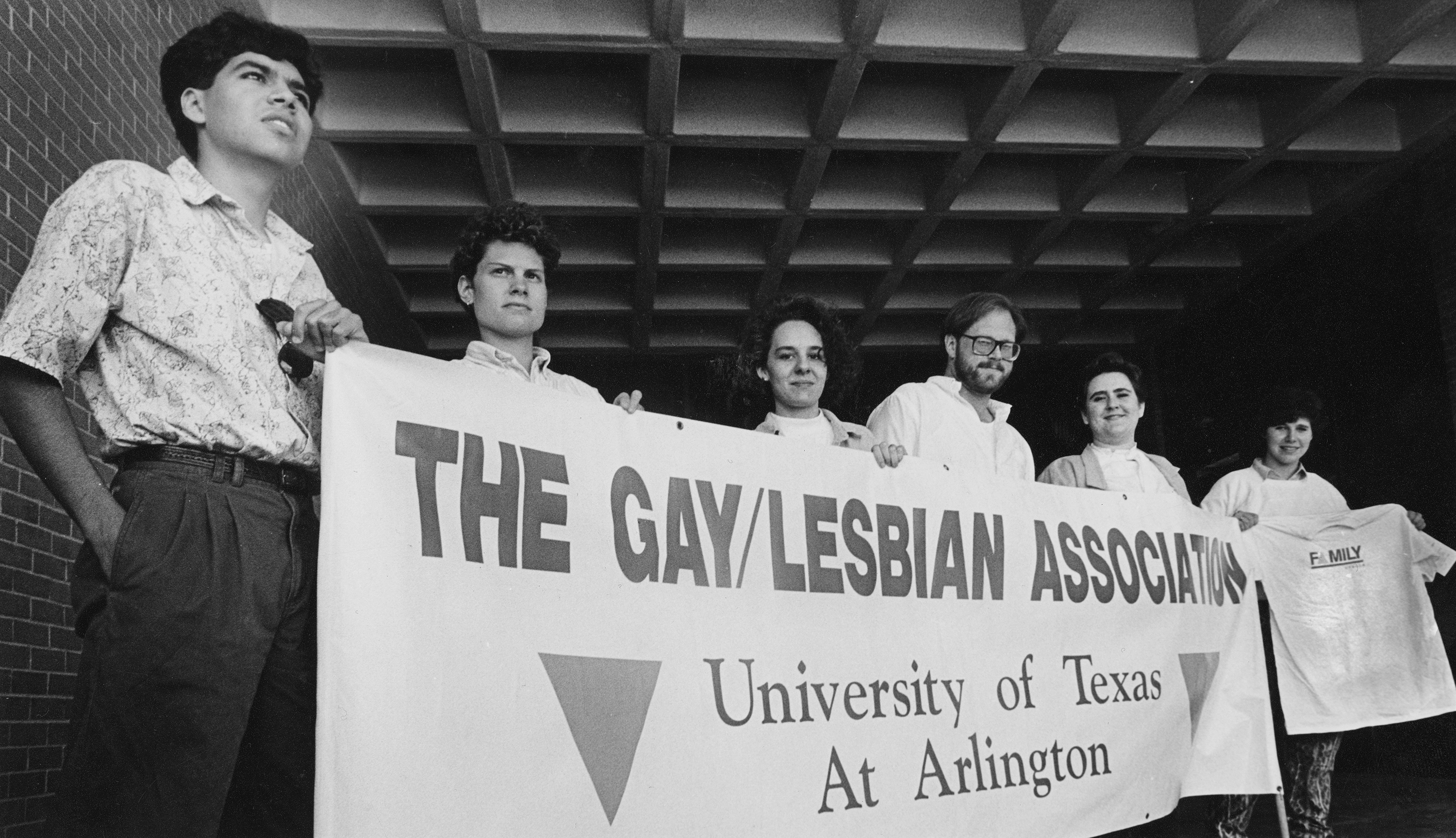 The Gay/Lesbian Association (GLA) of the University of Texas at Arlington members posed for a portrait, holding their banner. A member at the far right is holding a shirt reading "Family." The GLA was preparing to march in the upcoming "March on Austin for Lesbian-Gay Equal Rights," to be held on March 17, 1991, with their banner.
