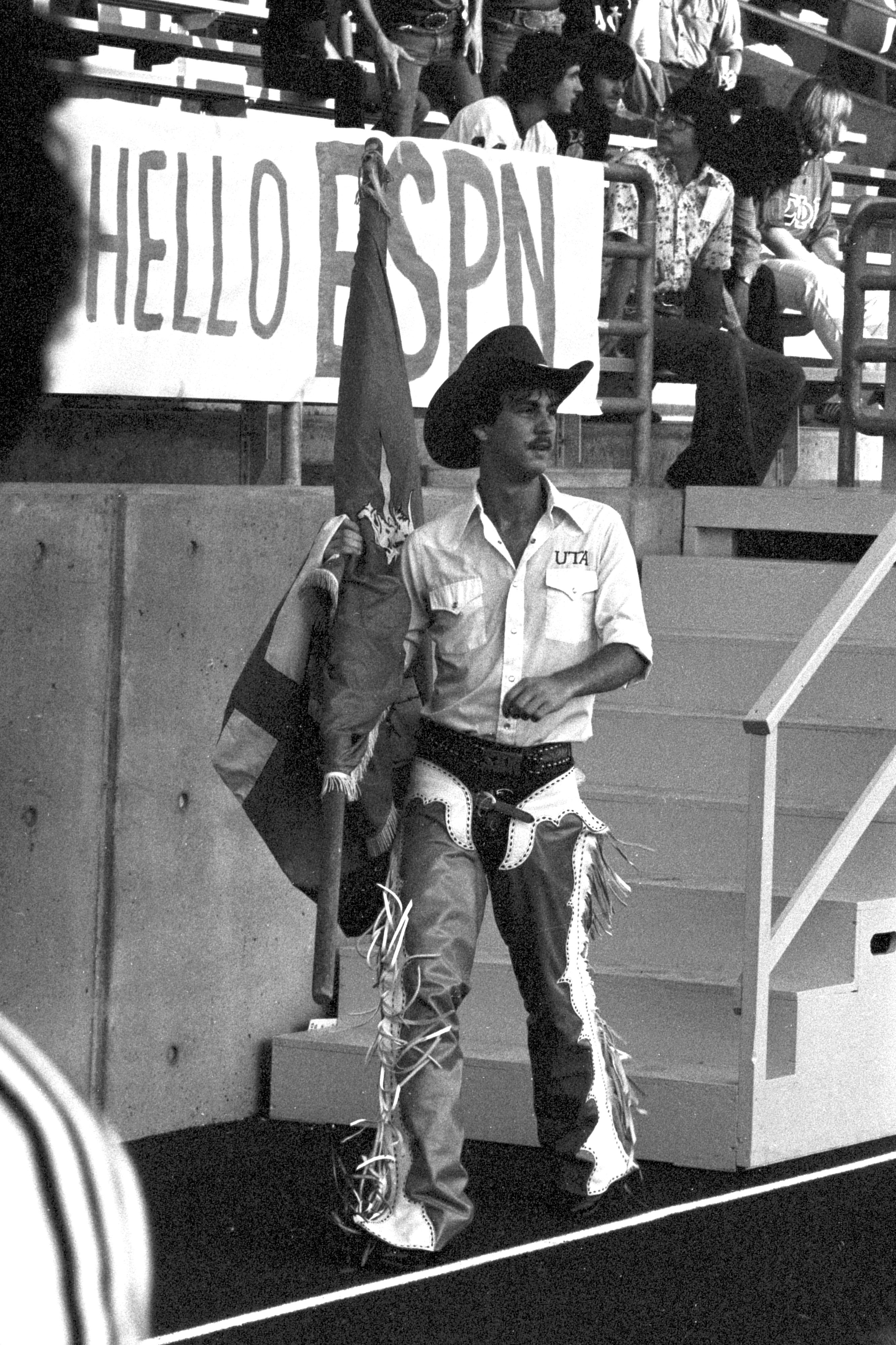 Man wearing white collared UTA shirt with cowboy hat holding a flag.