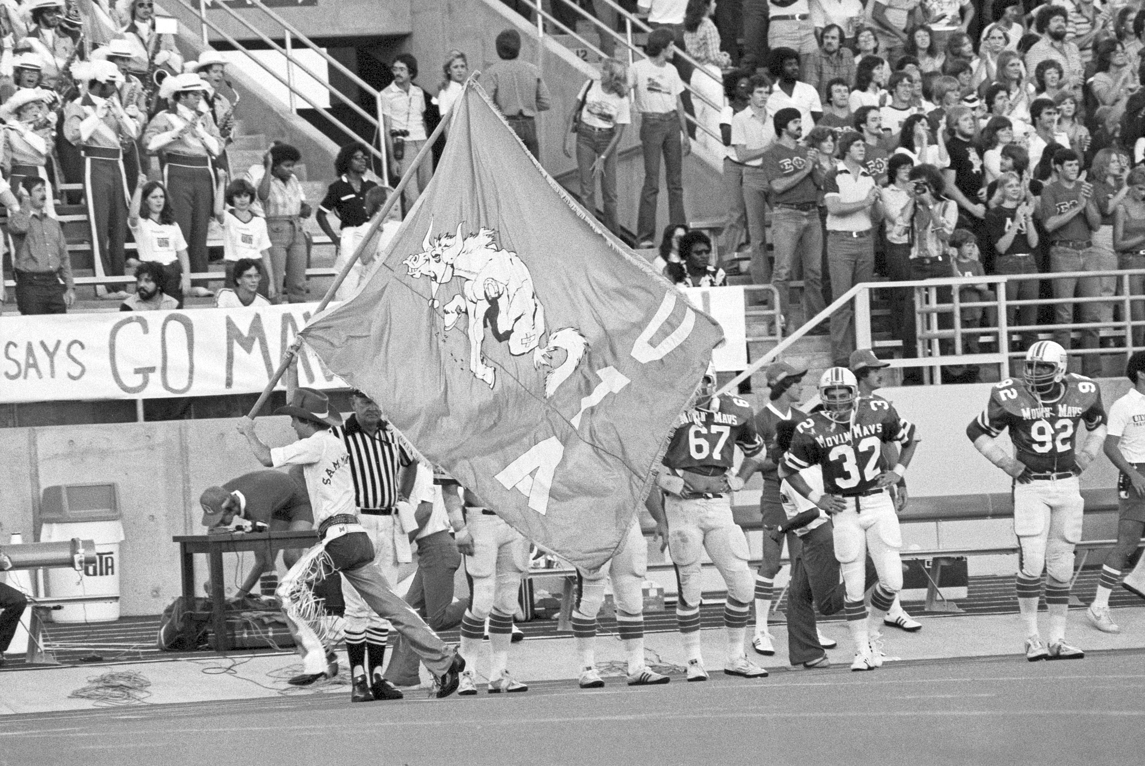 Man dressed in chaps and cowboy hat running with a large UTA flag while on the sidelines of a football game and a crowd is seen in the background.