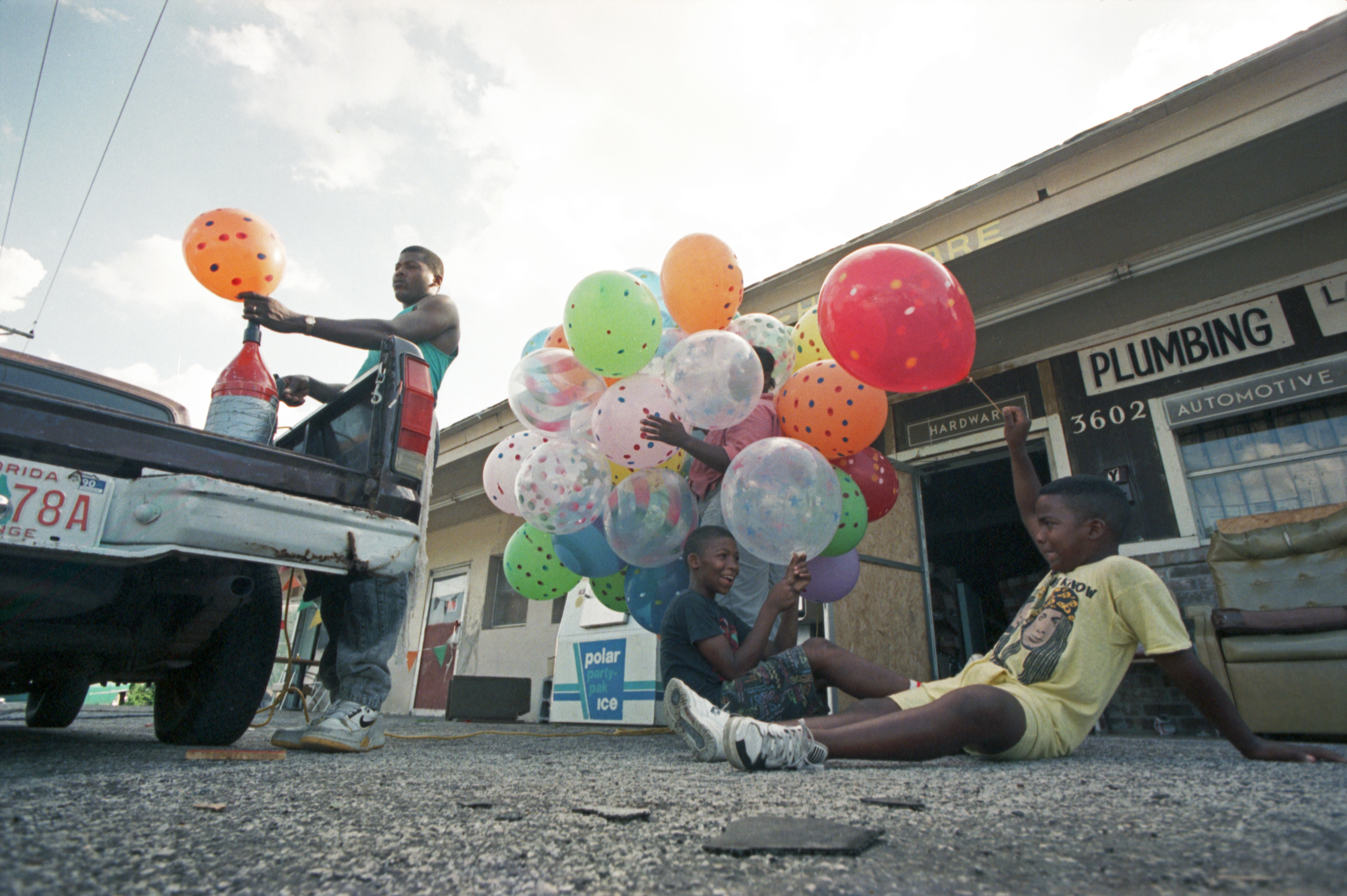 Color photo of three children with balloons and a man blowing up a balloon from a helium tank in the bed of a truck.