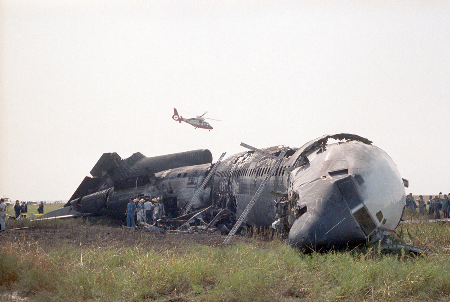 Color photo of a airplane crashed in a grassy field with a helicopter seen hovering above it and rescue workers seen gathered near the wreckage.