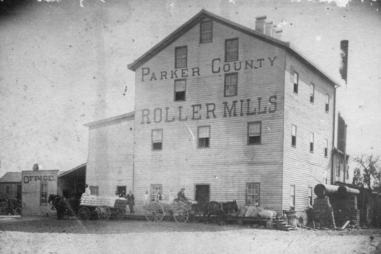 Photo of Parker County Roller Mills business, circa 1900. Shows a 3-story building with horses and horse carriages out front.