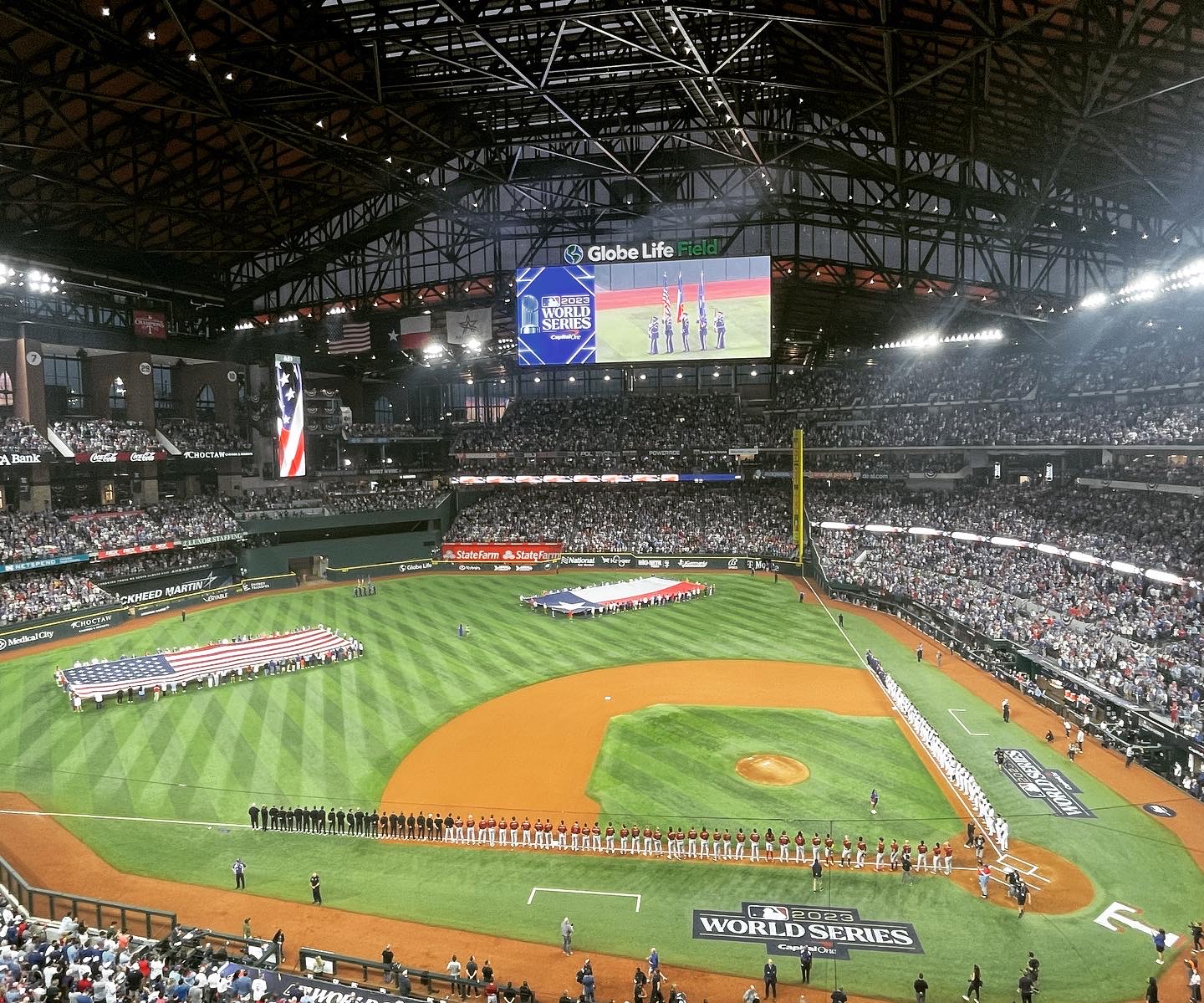 Image of Globe Life Field during game 2 of the 2023 World Series showing both teams lined up along the baselines and people on the field holding the American and Texas flags.