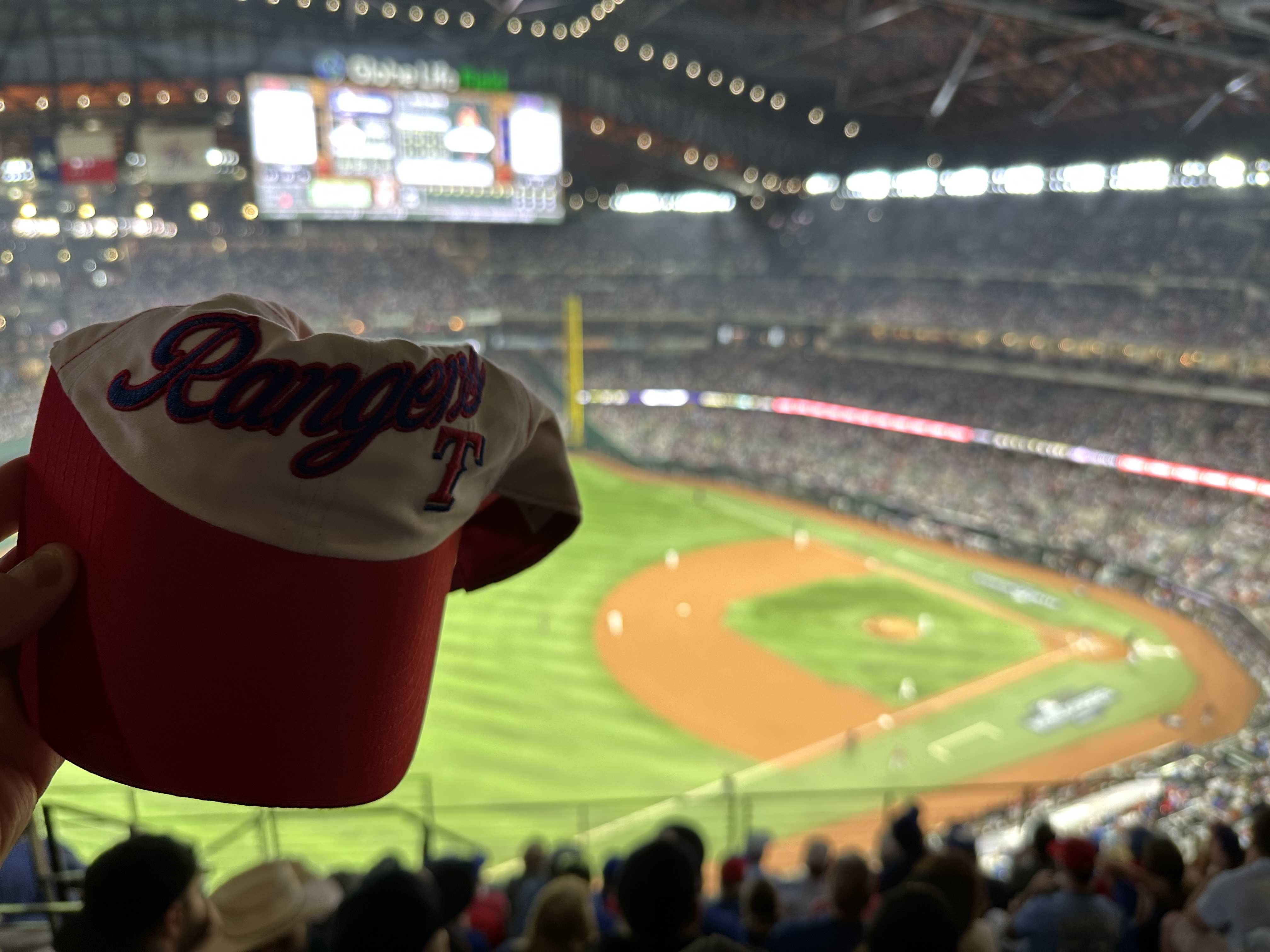 Image of someone holding a red and white Texas Rangers hat with Globe Life Field in the background. 
