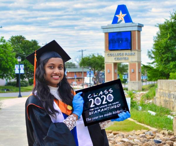 a student in graduation garb and latex gloves poses in front of college park center with a sign that reads: "Class 2020: The One Where We Were Quarantined"