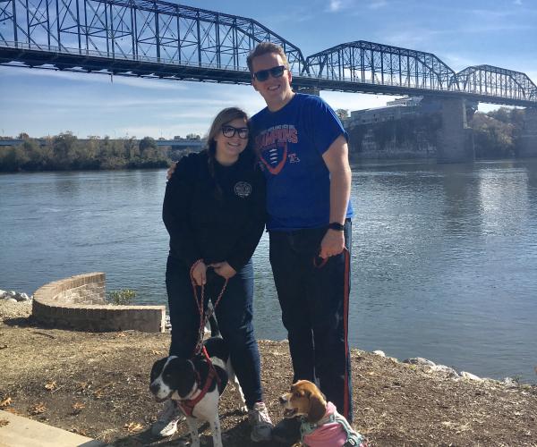 a young woman and young man pose in front of a bridge with their two dogs on leashes