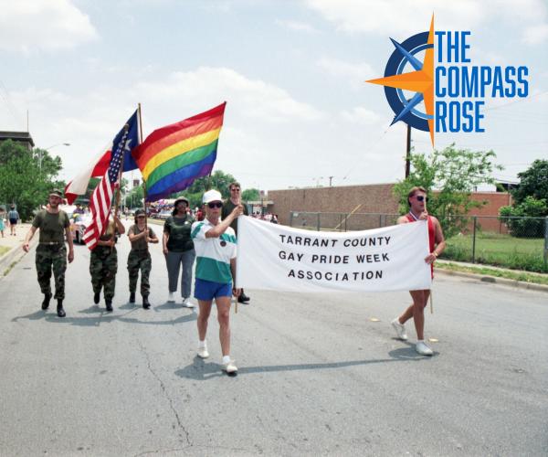 Members of the Tarrant County Gay Pride Week Association are seen carrying a banner along with three flags (the American flag, the Texas flag, and a rainbow flag representing gay pride) during the Gay Pride Week parade. The members carrying the flags are seen dressed in military-style camouflage clothing.