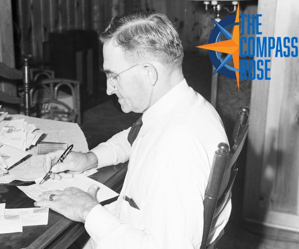 J.J. Boydstun at his table handwriting letters with a fountain pen. He is wearing a white shirt and tie, and has glasses.