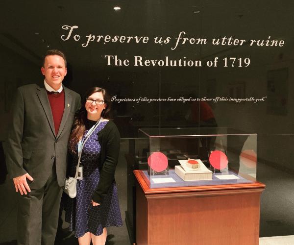 a white man and woman in nice clothes, standing in front of an exhibit display, which reads, "to preserve us from utter ruine: the revolution of 1719"