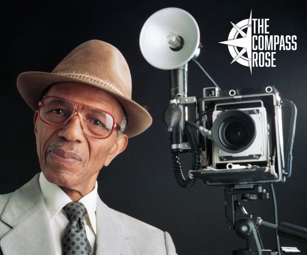 Portrait of Calvin Littlejohn posed next to a Graflex Crown Graphic camera with an external flash attached. 