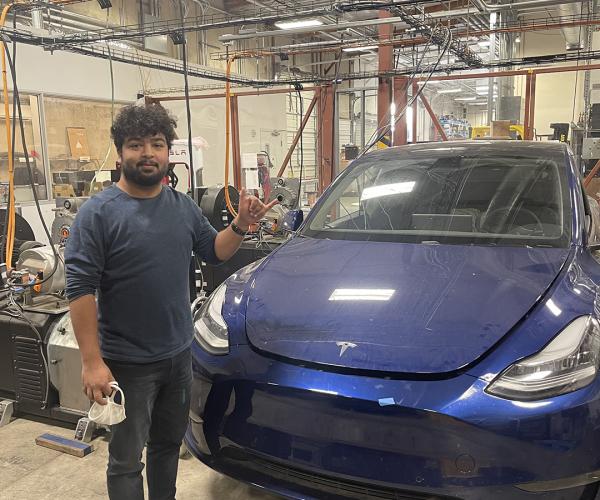 vikram gupta stands in a massive garage next to a blue model y tesla vehicle, which is a on a dyno machine for performance testing; vikram is giving the mav up hand gesture and smiling