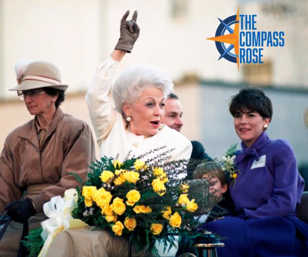 Ann Richards at her inauguration parade after being elected Governor of Texas. She is holding a bouquet of yellow roses and holding up her right gloved hand in a "Hook 'Em" gesture, while riding in a horse-drawn carriage with family members.