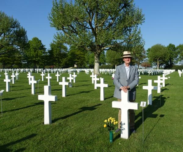 An older man in a gray suit jacket and beige brimmed hat stands with a cross adorned for Captain Chester Slaughter. Around and behind him stretches a large cemetery of identical crosses for other American soldiers who died overseas in World War two.