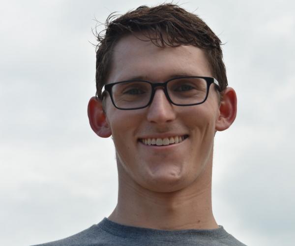 a young man in black glasses and a gray shirt smiles at the camera