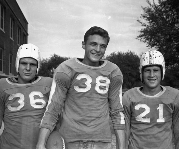 three young men stand side by side in their football uniforms, smiling for the camera; the two on the ends are wearing their helmets, while the middle man is not. behind them are trees and a brick building.