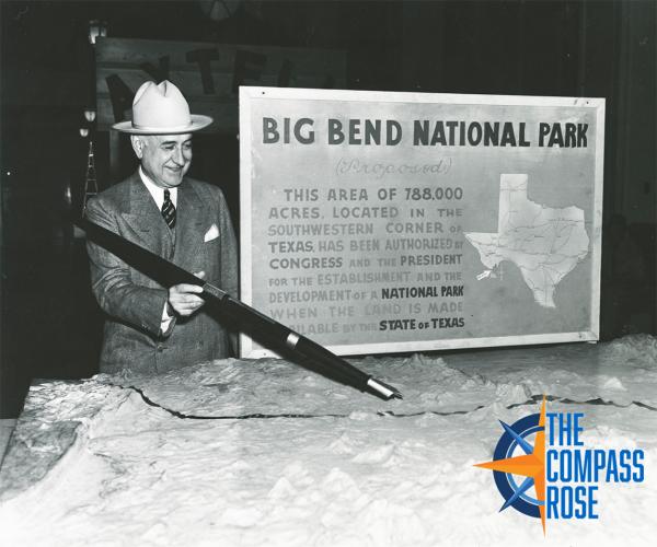 A man with a large pen points to a model of land with a sign to his left. The Compass Rose logo is in the lower right corner.
