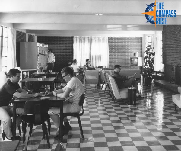 Male students in a dormitory common area, some at tables and others at couches. The building's interior brick walls and checkerboard-patterened flooring are clearly visible.