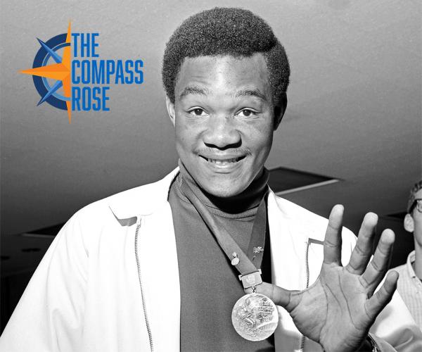 Black and white photograph of boxer George Foreman holding up an Olympic gold medal.