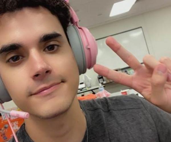 A young man wearing pink cat-ear headphone smiles at the camera, holding up two fingers in a peace symbol.
