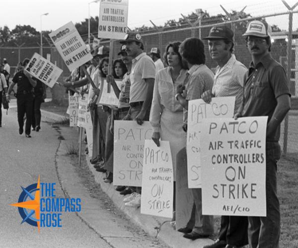 Group of strikers with picket signs standing in a picket line along a curb with a metal fence in the background. Orange and blue "The Compass Rose' logo in the bottom left corner.