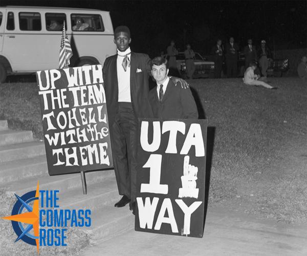 Two students standing together holding up signs protesting the Rebel theme on UTA's campus. One sign says "Up with the Team To Hell with the Theme." The other says "UTA 1 Way."
