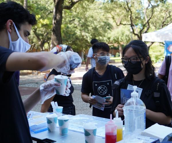 A young man pours purple syrup on a cup of ice while a student in a black mask looks on. Several students mill in a line behind them.