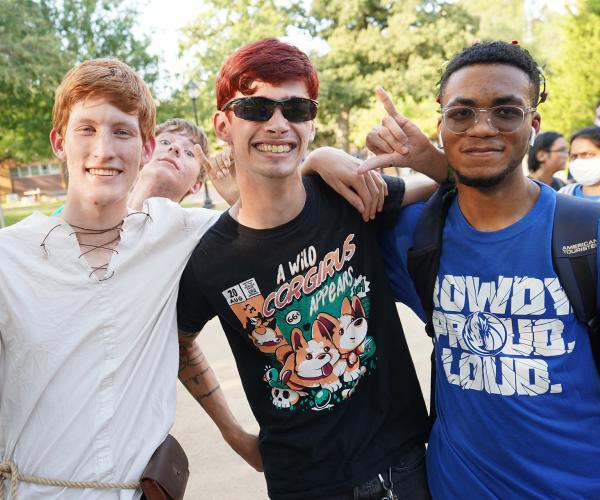 Three young men pose together in the Central Library Mall, giving the Mav Up hand gesture. One young man pokes his head behind the others.