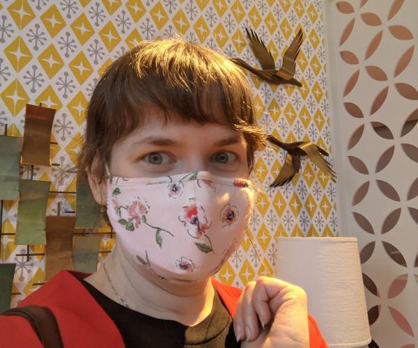 A woman with blunt bangs and a rose print face mask looks into the camera. The wallpaper behind her has an intense mod pattern.