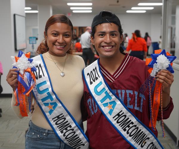 Two young people stand together wearing homecoming court sashes and hold up mums they have made.