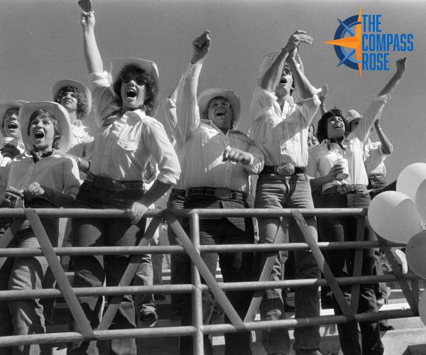 Spirit group cheering and ringing cowbells along the front rail of the stands at a football stadium, with "The Compass Rose" logo in the top right corner.