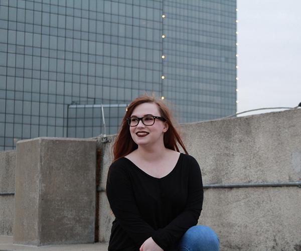 A young woman with long red hair and glasses smiles and looks up to the sky with an industrial background.