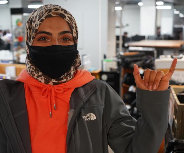A young woman wearing a leopard print headscarf and black face mask gives the Mav Up hand gesture in the U T A Central Library. She is wearing an orange hoodie underneath a gray The North Face jacket.