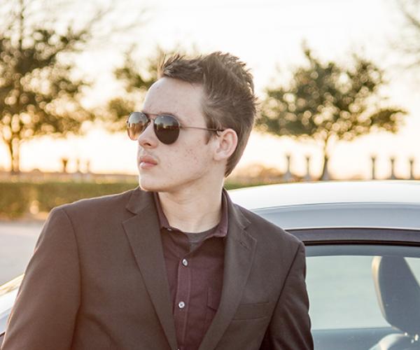 A man in a brown suit stands in front of a car. Aviator sunglasses frame his face, which is lit by warm sunshine.