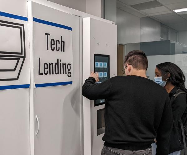 Two young people stand at a Tech Lending locker, navigating the user interface with their backs to the camera.