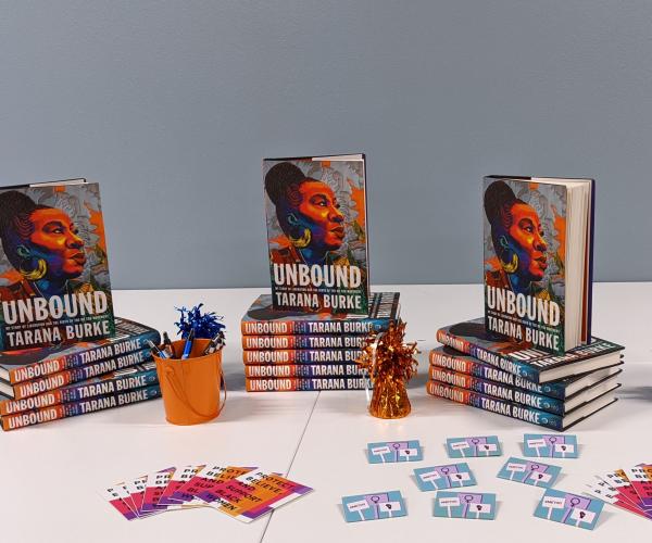 Three stacks of books are displayed on a table along with a bucket full of pens, some stickers, and some enamel pins.