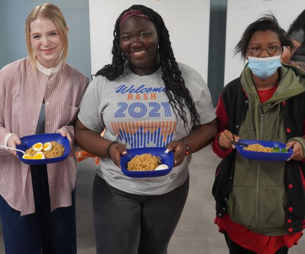 Three students stand together. They hold plastic blue plates which have ramen and soft-boiled eggs in them. 