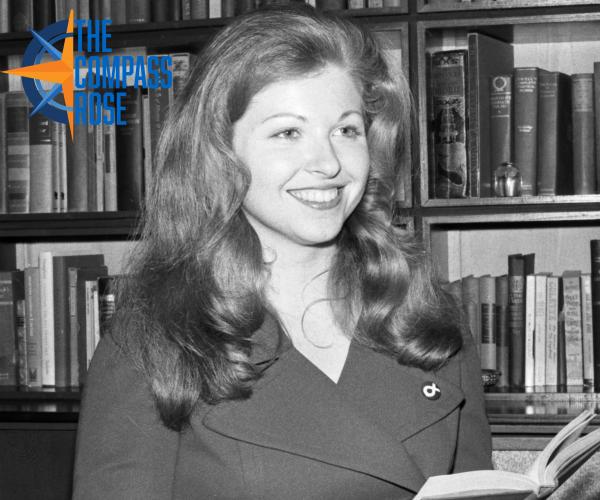 Woman smiling holding a book with bookshelves in the background. Logo with the words "The Compass Rose" in blue and orange is in the top left corner of the photo