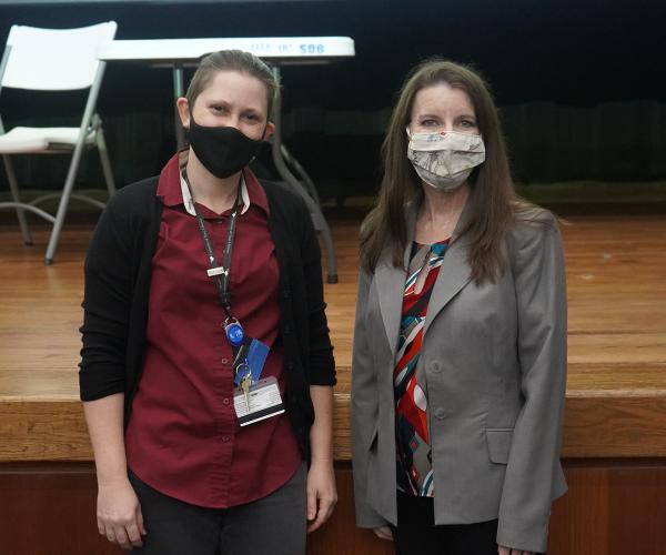 A nonbinary person in a maroon polo and woman in a gray tailored jacket, both wearing face masks, stand side by side in front of a wooden stage.