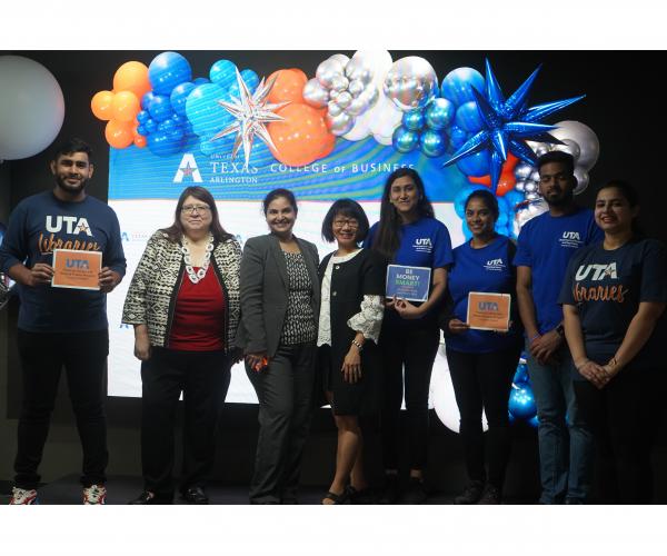 A group of people who hosted the Money Smart Workshop series stand in front of a display that reads, "University of Texas at Arlington, College of Business."