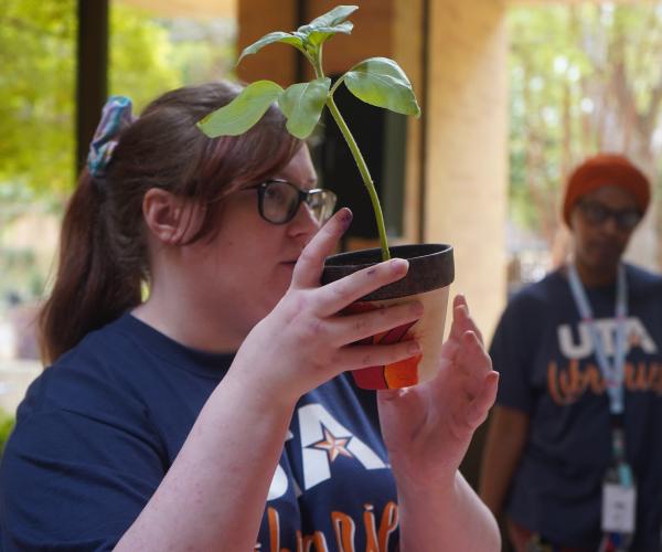 A woman in glasses holds a pot painted with white, orange, and red. A green plant sprouts from the pot, obscuring her face. 