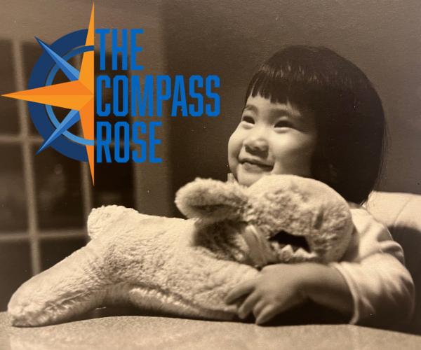 Picture of a Japanese American toddler smiling and looking towards the top left of the photo and holding a stuffed lamb, The Compass Rose logo at top left corner.
