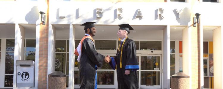 two students in graduation gowns shaking hands in front of the Central Library